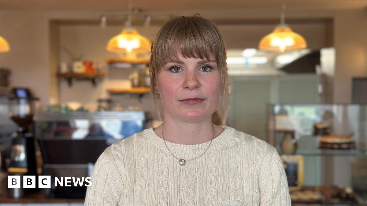 A woman with blonde hair and wearing a knitted jumper. She is standing in a cafe, the counter and some lighting is visible in the background.