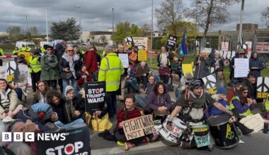 A group of about 50 people sit on the road holding signs with messages including Stop Trump's Wars, and No War on Iran. To their right is the base's wire fence.