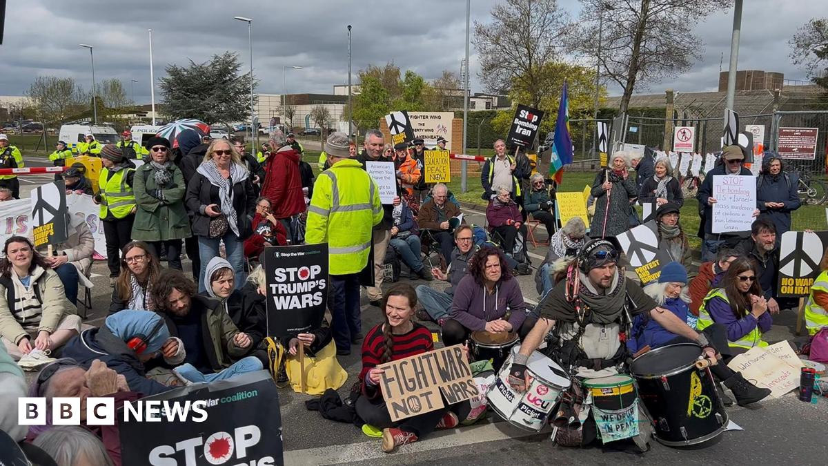 A group of about 50 people sit on the road holding signs with messages including Stop Trump's Wars, and No War on Iran. To their right is the base's wire fence.