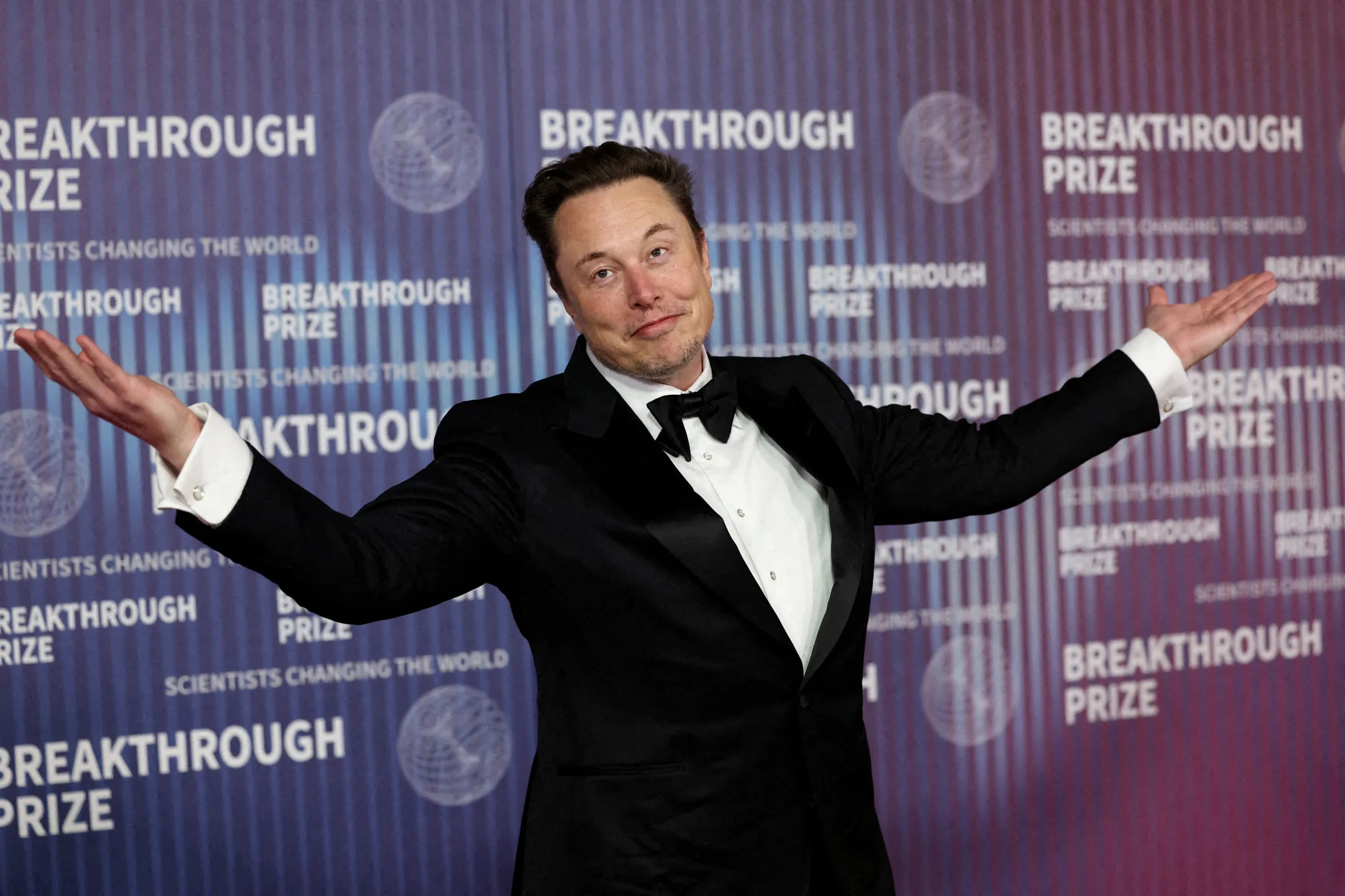 Elon Musk, in a tuxedo, shrugging with open arms on a red carpet with "Breakthrough Prize" repeated on the background.