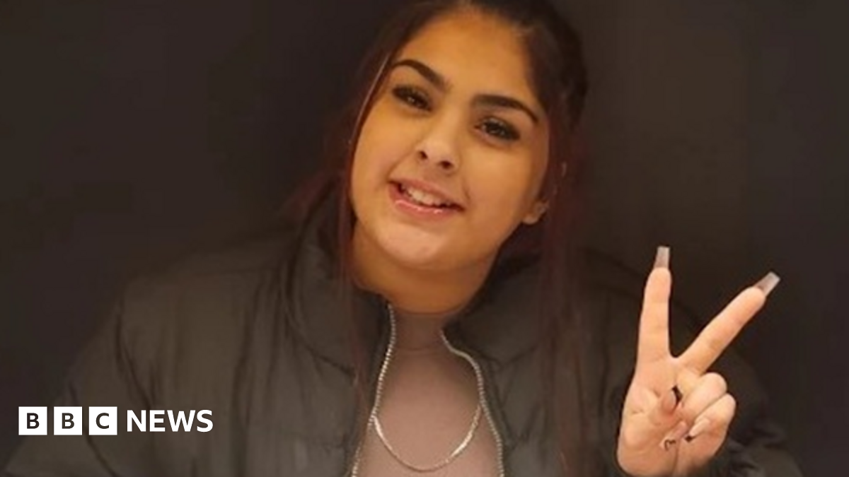 Aaliyah has dark hair in a ponytail, brown eyes and is smiling at the camera. She is wearing a dusty pink t-shirt with a black jacket over the top, she is holding up a peace sign with her fingers.