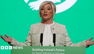 Michelle O'Neill standing at a green lecturn with a green background behind her. She is motioning with her hands. She is wearing a cream jacket and white shirt. She has blonde hair, tied back.