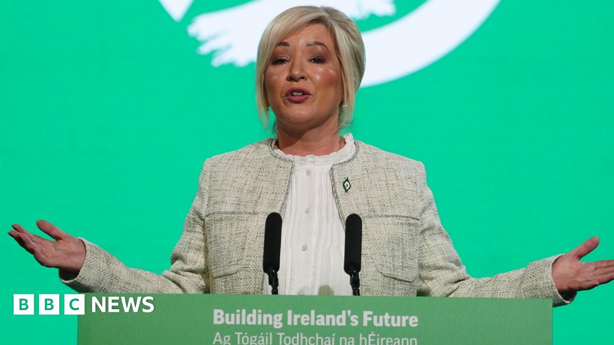 Michelle O'Neill standing at a green lecturn with a green background behind her. She is motioning with her hands. She is wearing a cream jacket and white shirt. She has blonde hair, tied back.