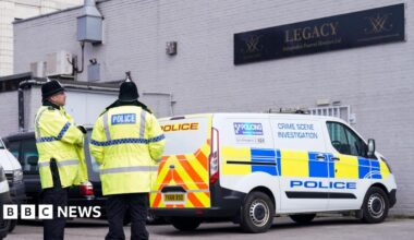 Two police officers wearing yellow jackets and black helmets stand outside a white bricked building. A crime scene investigation police van is parked underneath a black sign that reads Legacy Independent Funeral Directors