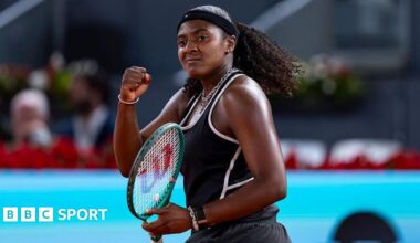 Hailey Baptiste, wearing a black vest and shorts, black headband and a ponytail, holds her green Wilson racket in her left hand and punches her right fist in celebration
