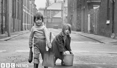 A black and white image shows two children in an empty terraced street. One is carrying a shovel and brush the other is bent over lifting a bucket. The first has check trousers tucked into wellies and a thick jumper which looks a little too big. The other wears a long, warm check coat. Both are looking into the camera.