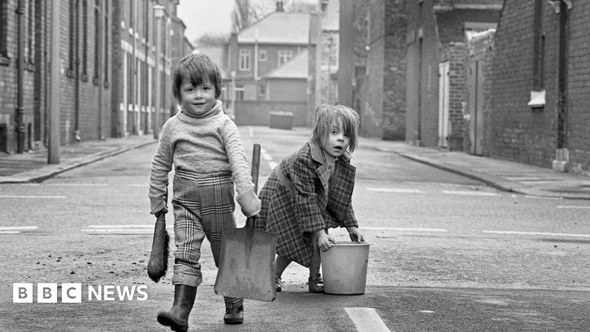 A black and white image shows two children in an empty terraced street. One is carrying a shovel and brush the other is bent over lifting a bucket. The first has check trousers tucked into wellies and a thick jumper which looks a little too big. The other wears a long, warm check coat. Both are looking into the camera.