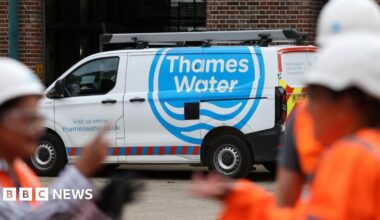 Blurry people in hi-vis safety gear and white helmets stand in the foreground, in front of a white van with a blue Thames Water logo.
