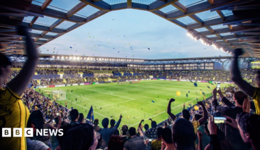 A CGI image of fans in the ground at the Triangle celebrating with many fans cheering and blue and yellow tape flying around in the air.