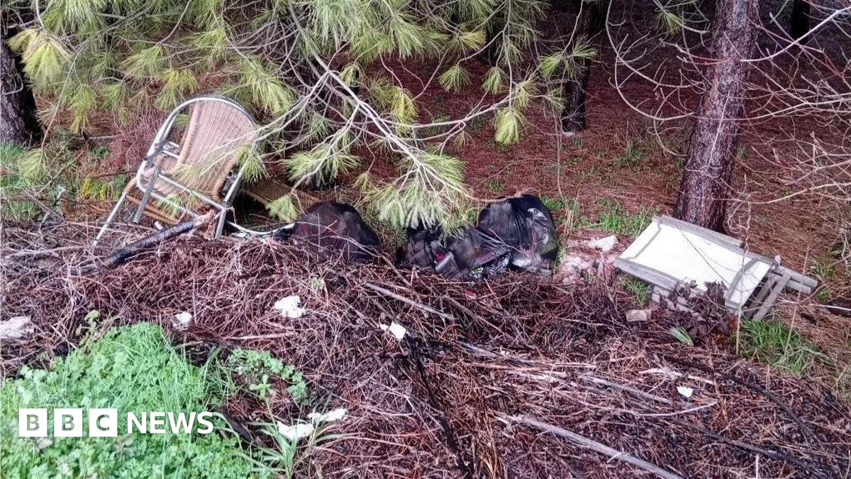 A haul of rubbish dumped at the bottom of a slope in a wooded area, There are wicker chairs with metal arms, a foldable white table and multiple black bin bags of household rubbish.