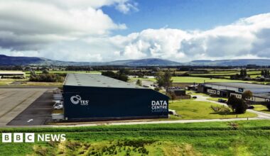A aerial view of a warehouse type building that is navy. It says "data centre" in white writing along the side. Behind it are rolling hills set against a sky with some blue and some clouds. the building is in a semi-rural area surrounded by green fields with one other smaller white industrial building to the right.