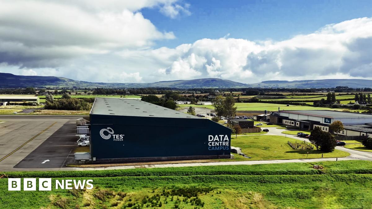 A aerial view of a warehouse type building that is navy. It says "data centre" in white writing along the side. Behind it are rolling hills set against a sky with some blue and some clouds. the building is in a semi-rural area surrounded by green fields with one other smaller white industrial building to the right.