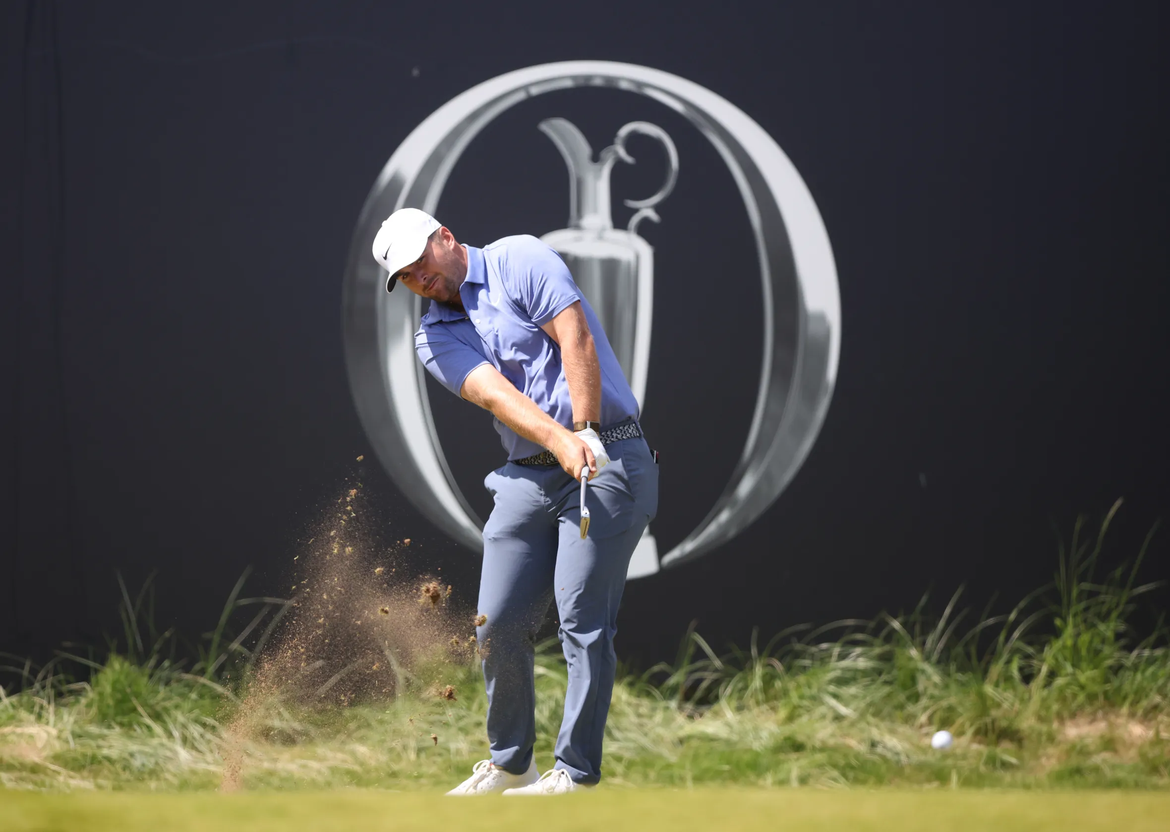 Golfer Chris Gotterup hitting a golf ball with dirt flying up from the swing, against a black wall with the Open Championship logo.