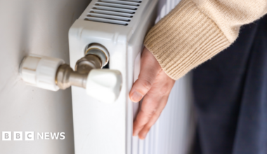 Someone is holding their hand against a radiator. Their face cannot be seen. The radiator is white and the person is wearing a yellow jumper and navy bottoms.