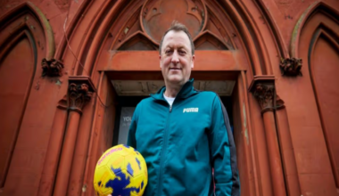Tony stands outside his community hub in Birkenhead with a football in hand. He is smiling and wears a teal green matching tracksuit.