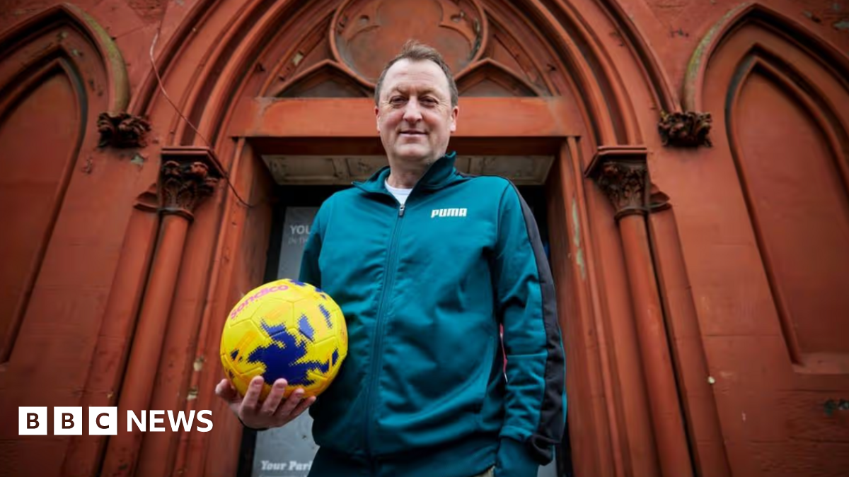 Tony stands outside his community hub in Birkenhead with a football in hand. He is smiling and wears a teal green matching tracksuit.