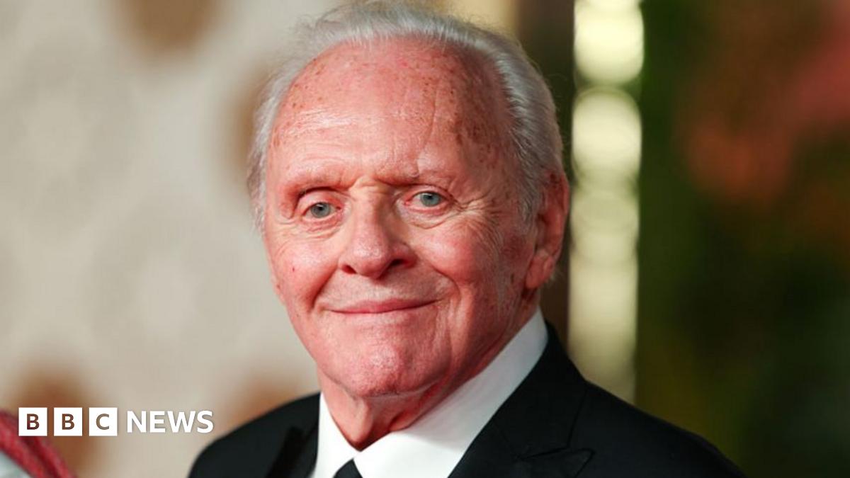 Sir Anthony Hopkins is stood by a Range Rover wearing a brown suit jacket and trousers, and a hat. He is surrounded by people from a production company for his new film.