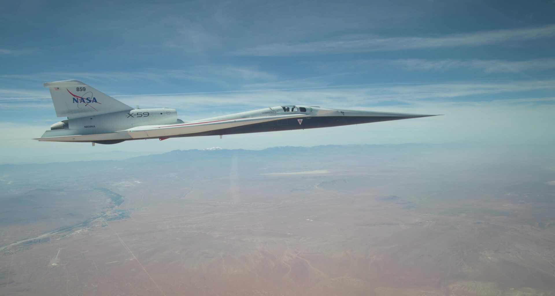 NASA’s X-59 quiet supersonic research aircraft flies above the Mojave Desert on a clear day. The aircraft is white with light gray, red, and blue accent colors. A NASA logo is visible on its tail, along with the number 859 above it. It appears to be flying level over the desert land-scape, and in the distance, you can see a mountain range on the horizon.
