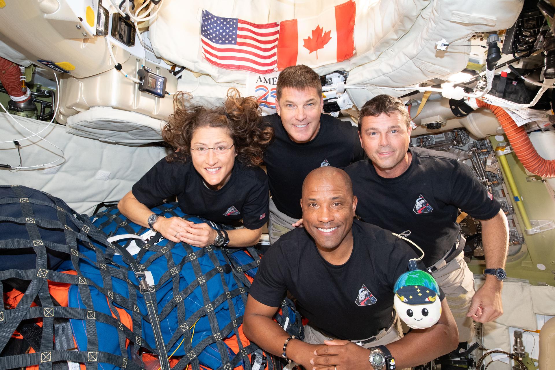 The crew of Artemis II—four astronauts—floats inside their Orion spacecraft. American and Canadian flags are visible in the background, in the top center of the frame. A zero gravity indicator—a smiling, plush depiction of the Moon and Earth—floats with the astronauts.