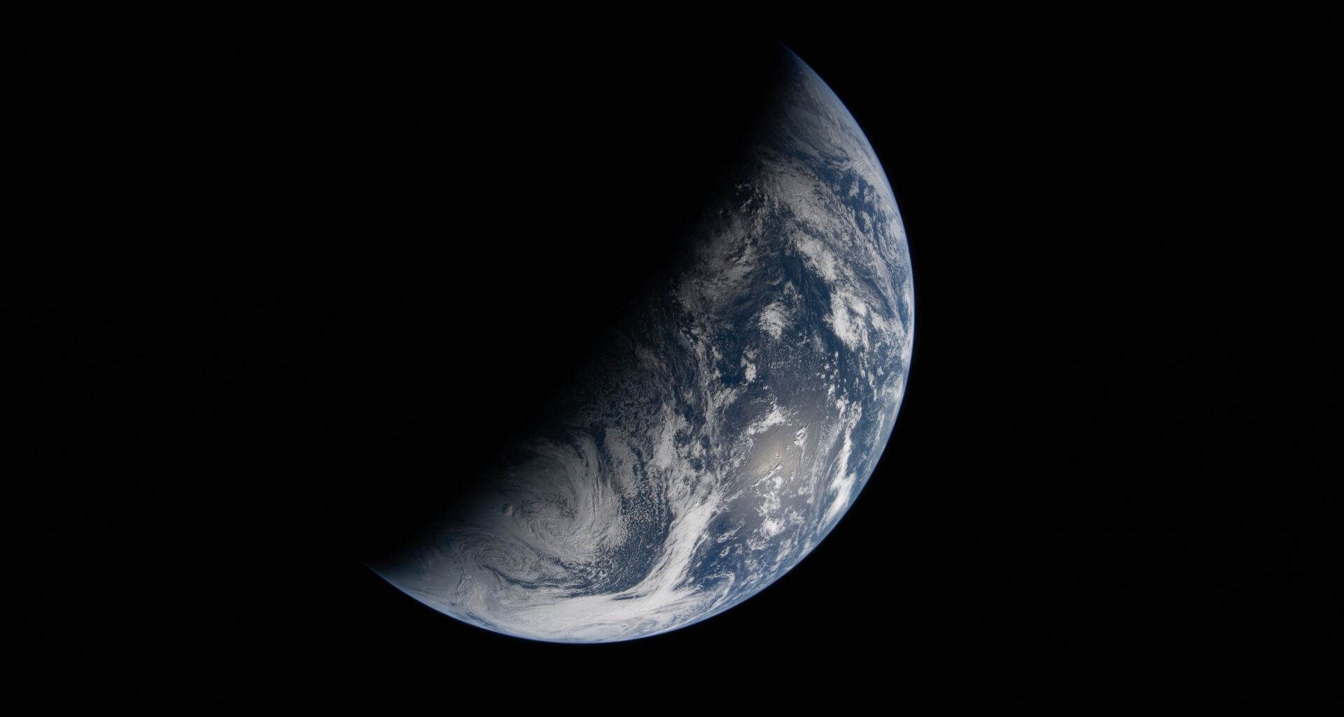 Image of Earth split by the terminator line. The terminator divides Earth’s surface diagonally, shrouding the top left half in darkness. Earth’s illuminated surface is mostly blue and covered in swirling white clouds.