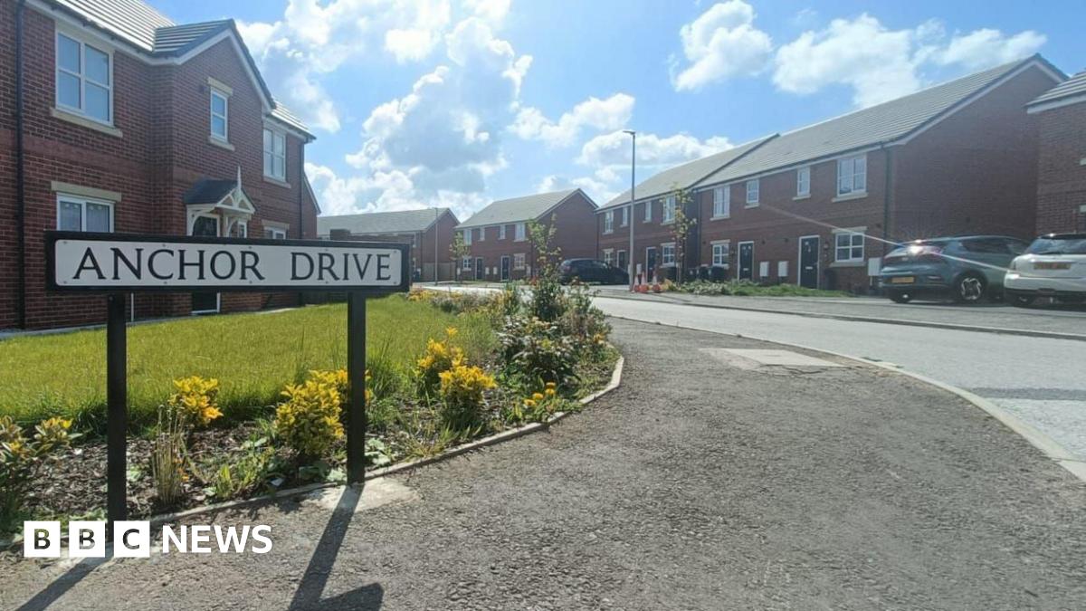 A sign reads Anchor Drive as the road leads round into a residential area - with houses on both sides of the road and cars in the drive