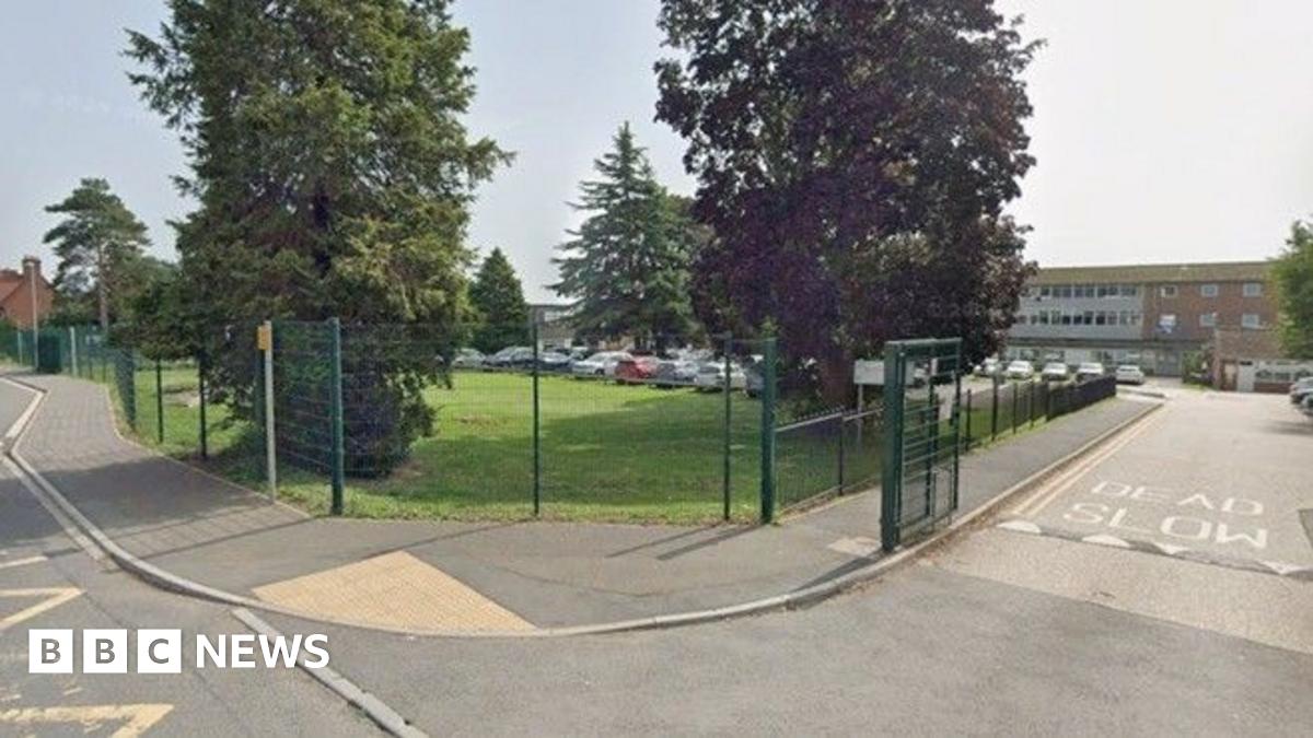 Google streetview image of Westfield Academy school. The school buildings are located behind a car park, with trees and grass in the foreground, and a green metal fence.
