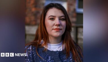 A still image of Jodie Morrow. She is staring at the camera, while she is stood against a wall outside. She has long, light coloured hair and is wearing a blue, knitted cardigan over a white t-shirt.