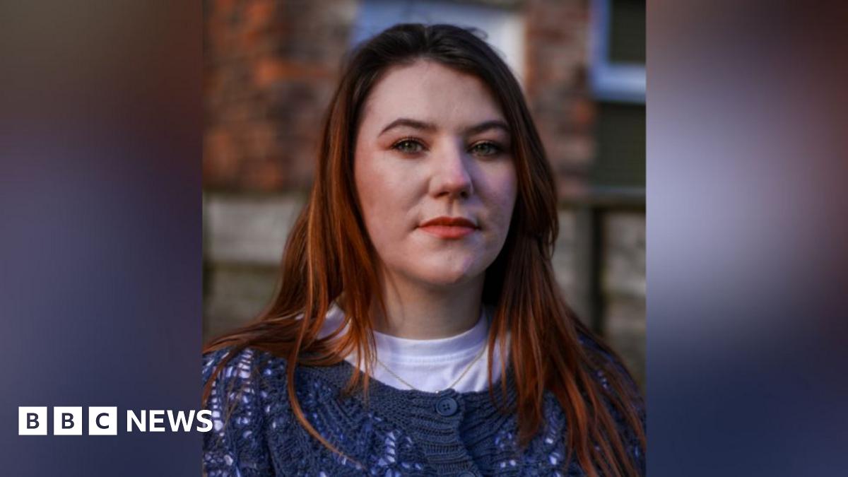 A still image of Jodie Morrow. She is staring at the camera, while she is stood against a wall outside. She has long, light coloured hair and is wearing a blue, knitted cardigan over a white t-shirt.