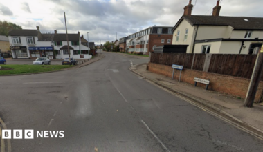 Greenfield Road leading into Flitwick, with houses and small shops on either side of the road and double yellow lines.