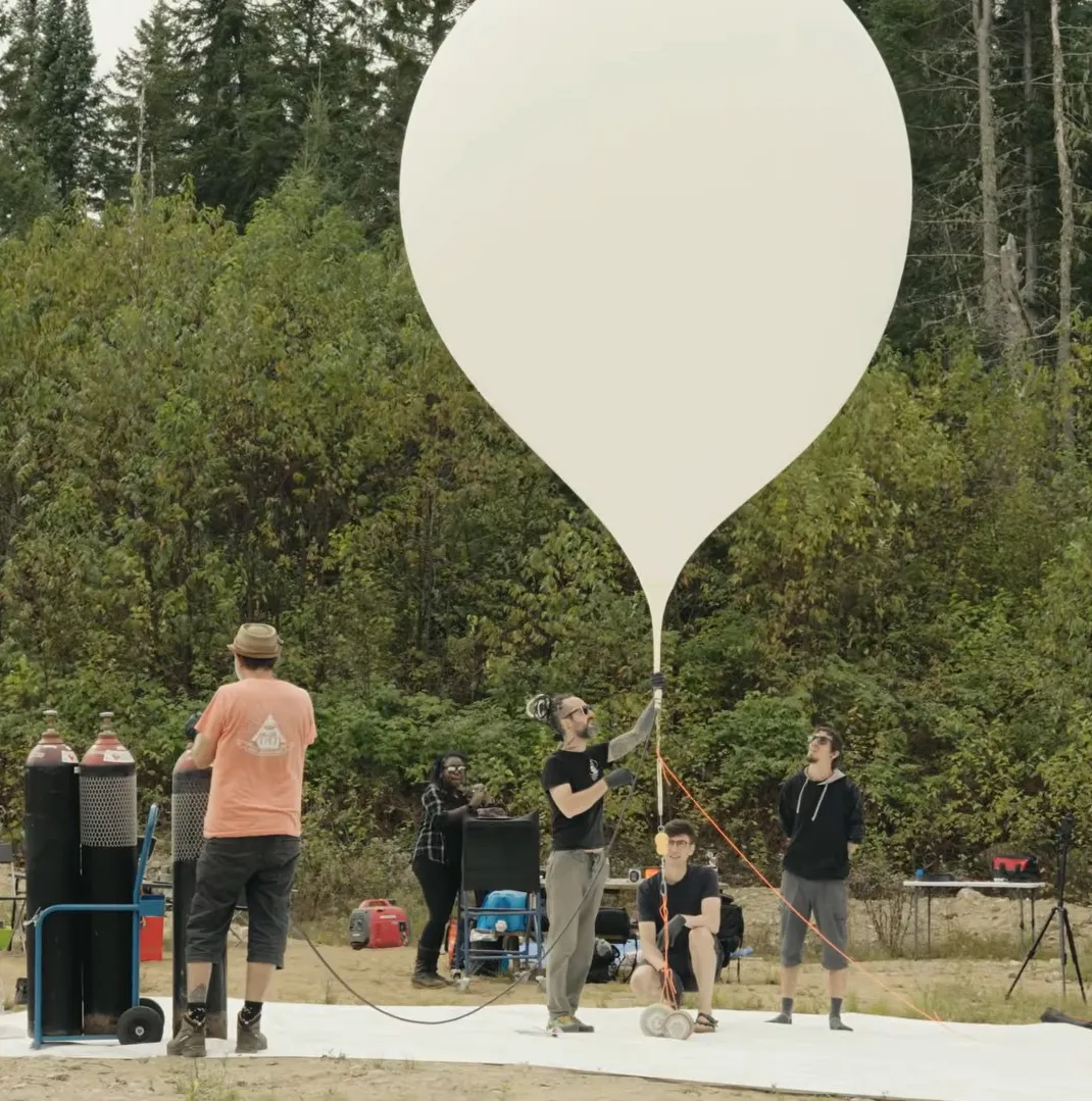 Five people preparing a large white balloon for launch in a forested area.