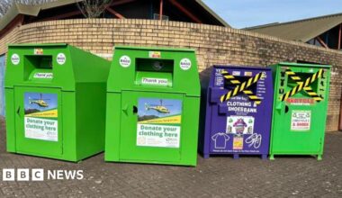 Image of four clothes and shoes recycling banks, lined up against a brick wall. In the background can be seen the roof of a building.