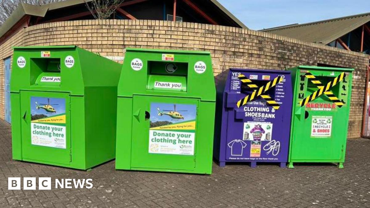 Image of four clothes and shoes recycling banks, lined up against a brick wall. In the background can be seen the roof of a building.
