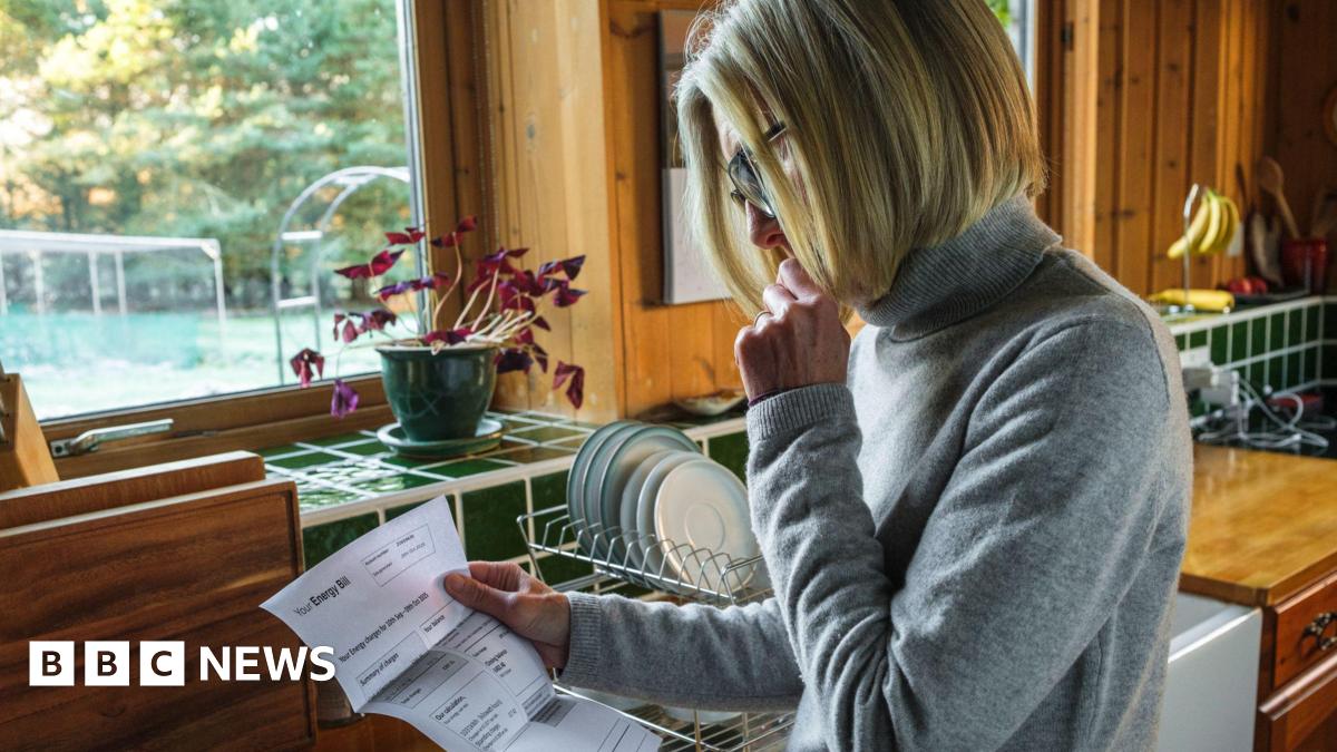 Woman looks at a bill in her kitchen with plates on the draining board behind her.