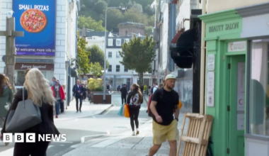 The image shows St Helier high street on a dry day. Members of the public are walking along the street to visit the shops.