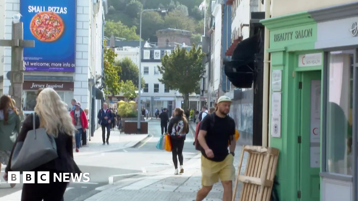 The image shows St Helier high street on a dry day. Members of the public are walking along the street to visit the shops.