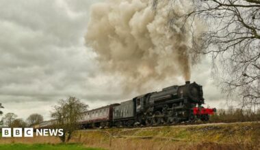 An old train on the Churnet Valley Railway emits steam as it travels along the track through the countryside.