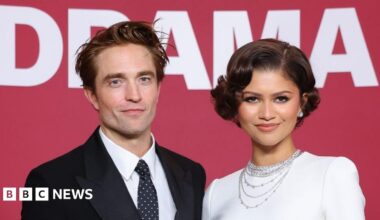 Robert Pattinson and Zendaya smiling at the camera. He has brown hair and is wearing a black suit jacket, black tie with white polka dots and a white shirt. Zendaya is wearing a white dress and diamond earrings and necklaces. She has short brown hair.