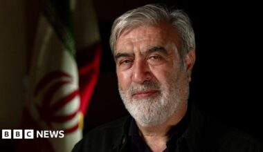 A man with a grey beard and hair looks into the camera. He is wearing a black shirt and behind him is the Iranian flag.