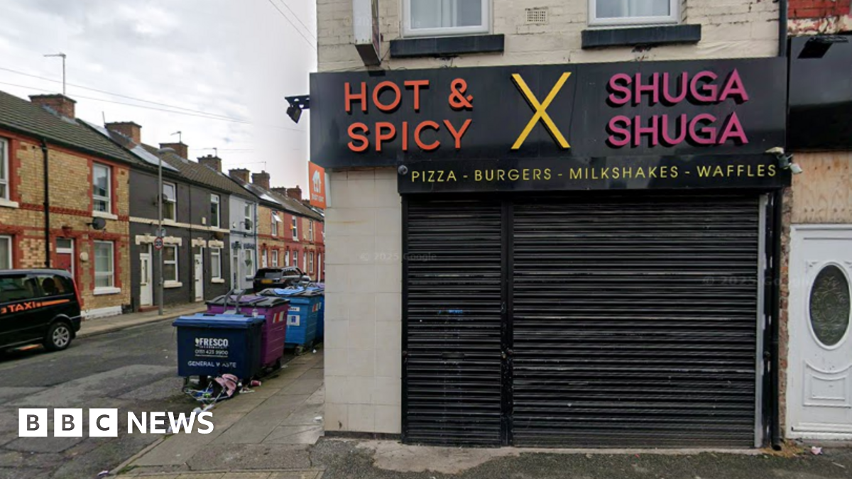 A shop has a sign saying 'Hot and Spicy', 'X' and 'Shuga Shuga' with 'pizzas - burgers - milkshake - waffles' underneath. Black shutters are down across the shop front. Industrial waste bins can be seen in a side street.