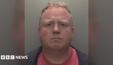 Robert Bush with short, light brown hair is shown in a straight‑on mugshot photograph. He has a neutral expression and is wearing a dark jacket over a red shirt. The background is plain and grey, and a Humberside Police badge watermark appears in the upper right corner of the image.