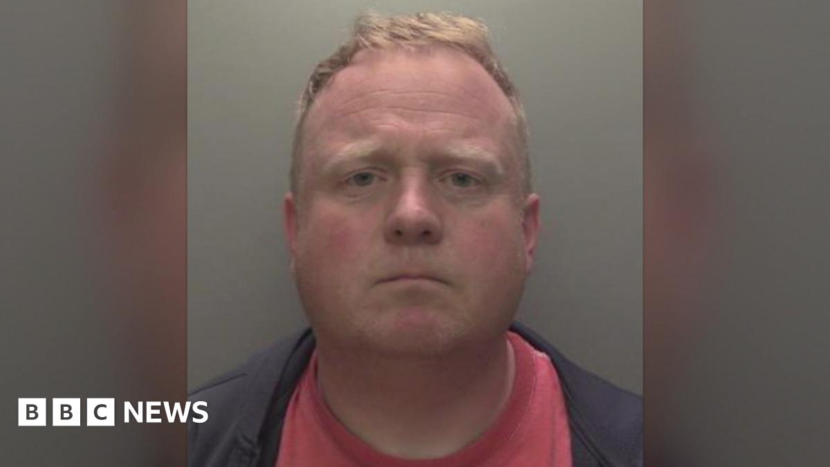 Robert Bush with short, light brown hair is shown in a straight‑on mugshot photograph. He has a neutral expression and is wearing a dark jacket over a red shirt. The background is plain and grey, and a Humberside Police badge watermark appears in the upper right corner of the image.