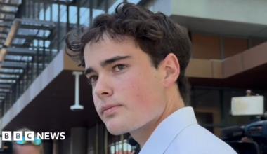 A young man with brown hair and a white shirt looks at the camera