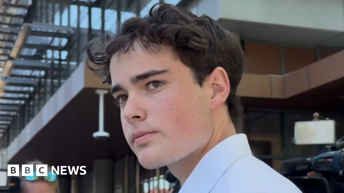 A young man with brown hair and a white shirt looks at the camera