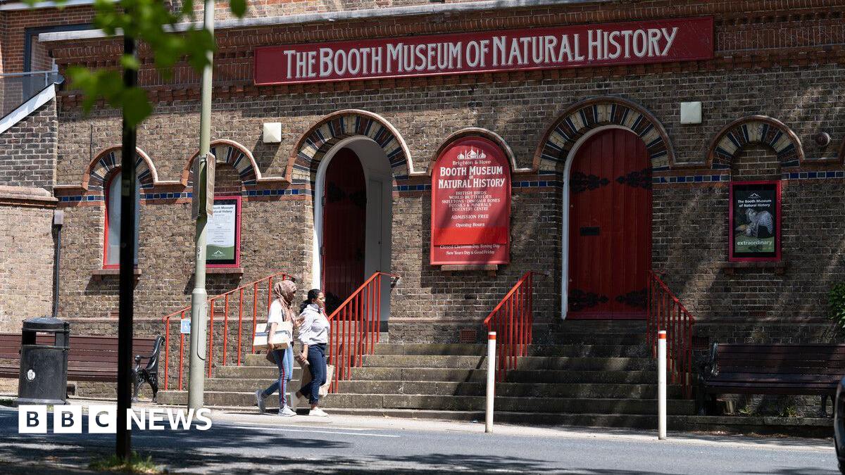 The building of The Booth Museum of Natural History. In front of the building there are red stair railings and two people walking past the building.