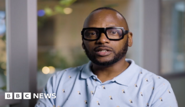 Christopher Joell-Deshields looks at the camera seriously, he is wearing a baby blue polo shirt with tiny puffins on it. His glasses are thick-rimmed black glasses and his hair is cropped short.
