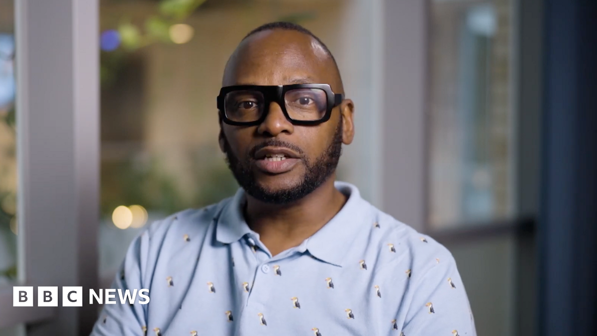 Christopher Joell-Deshields looks at the camera seriously, he is wearing a baby blue polo shirt with tiny puffins on it. His glasses are thick-rimmed black glasses and his hair is cropped short.