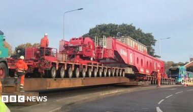 A general view of an abnormal load on a road. Highways workers in orange hi-vis  coats and trousers stand near or on the abnormal load.