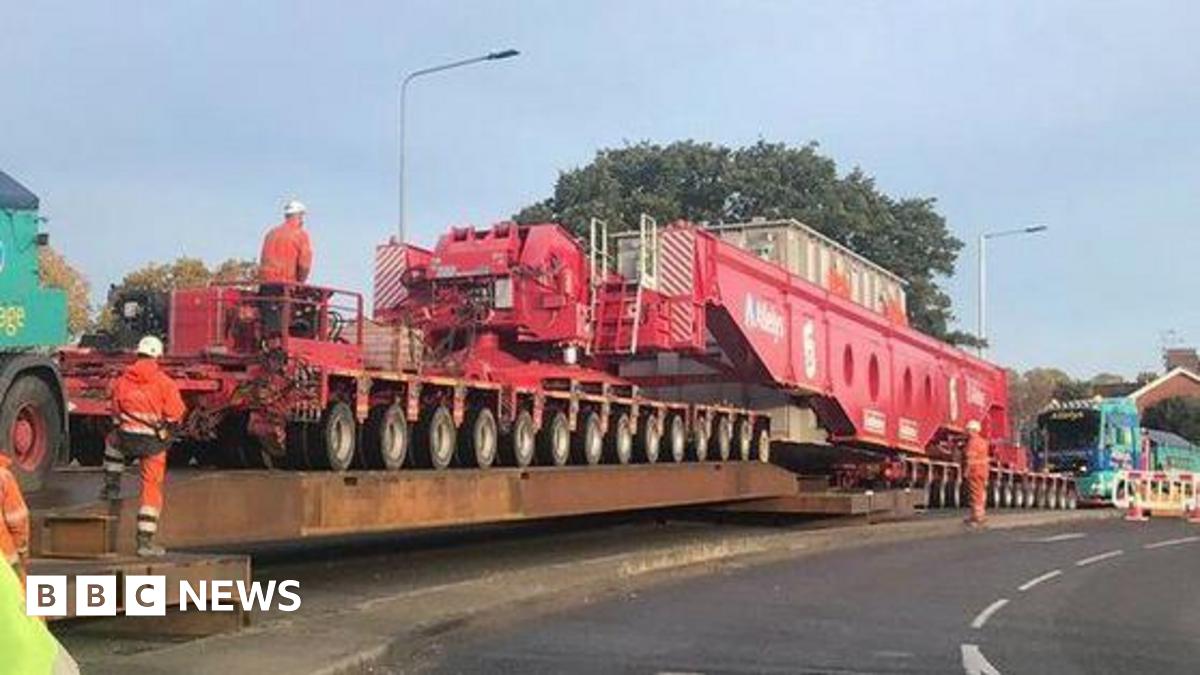 A general view of an abnormal load on a road. Highways workers in orange hi-vis  coats and trousers stand near or on the abnormal load.