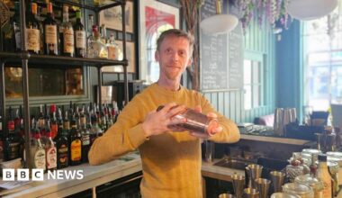 A blond man in a yellow jumper is holding a cocktail shaker. He is standing inside a bar.