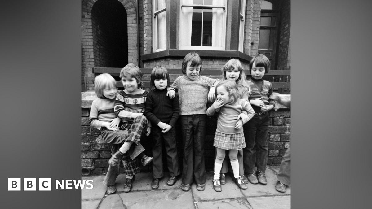 Black and white photograph of seven young children aged three to seven, in typical 1970s clothing, stood on a residential street with a Victorian house behind